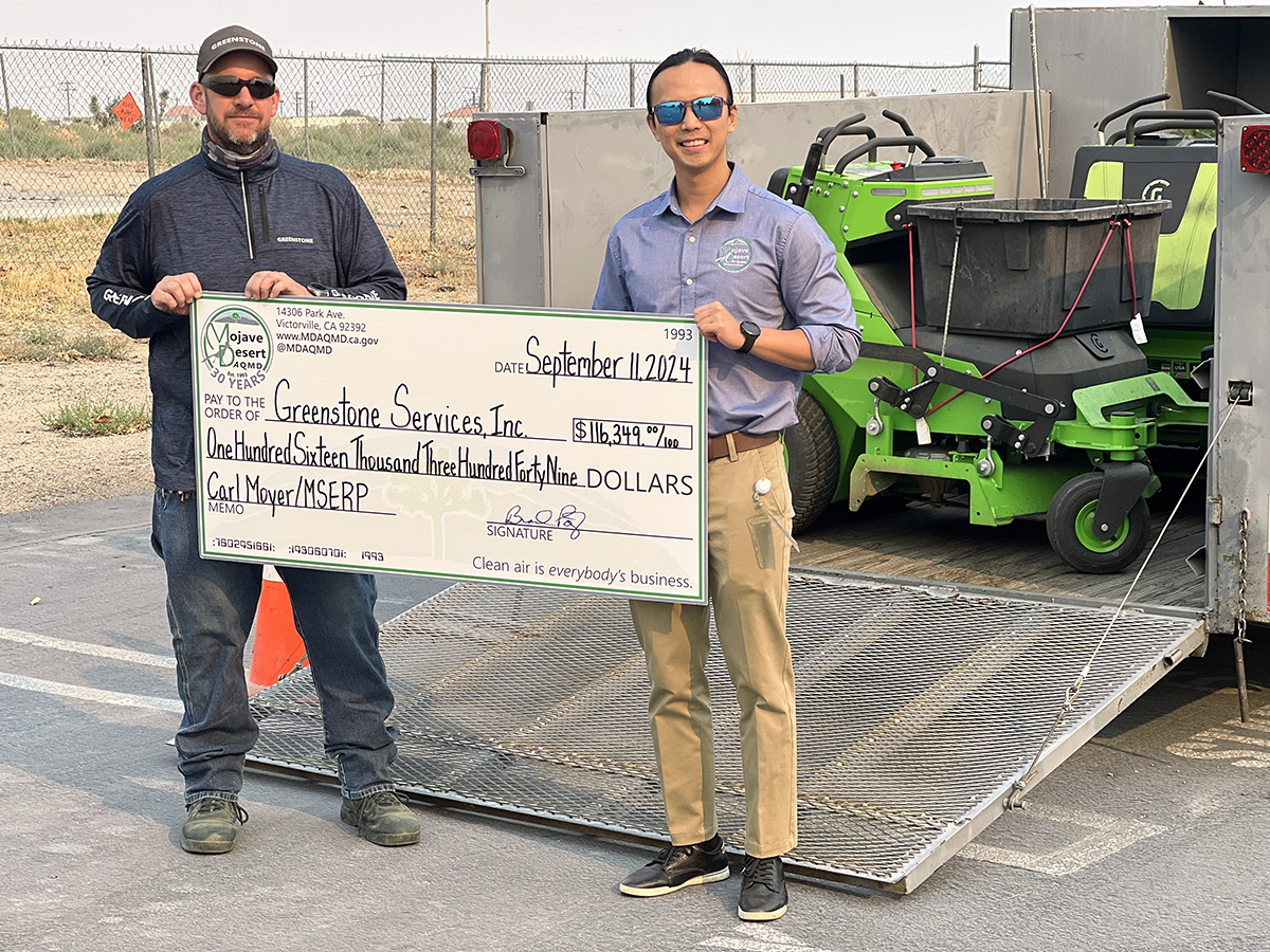 Photo shows two people holding a large foam-board check with landscaping equipment in the background.